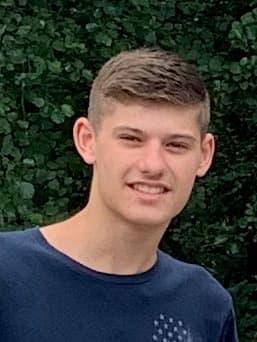 Smiling young man with short brown hair wearing a blue shirt against green foliage.