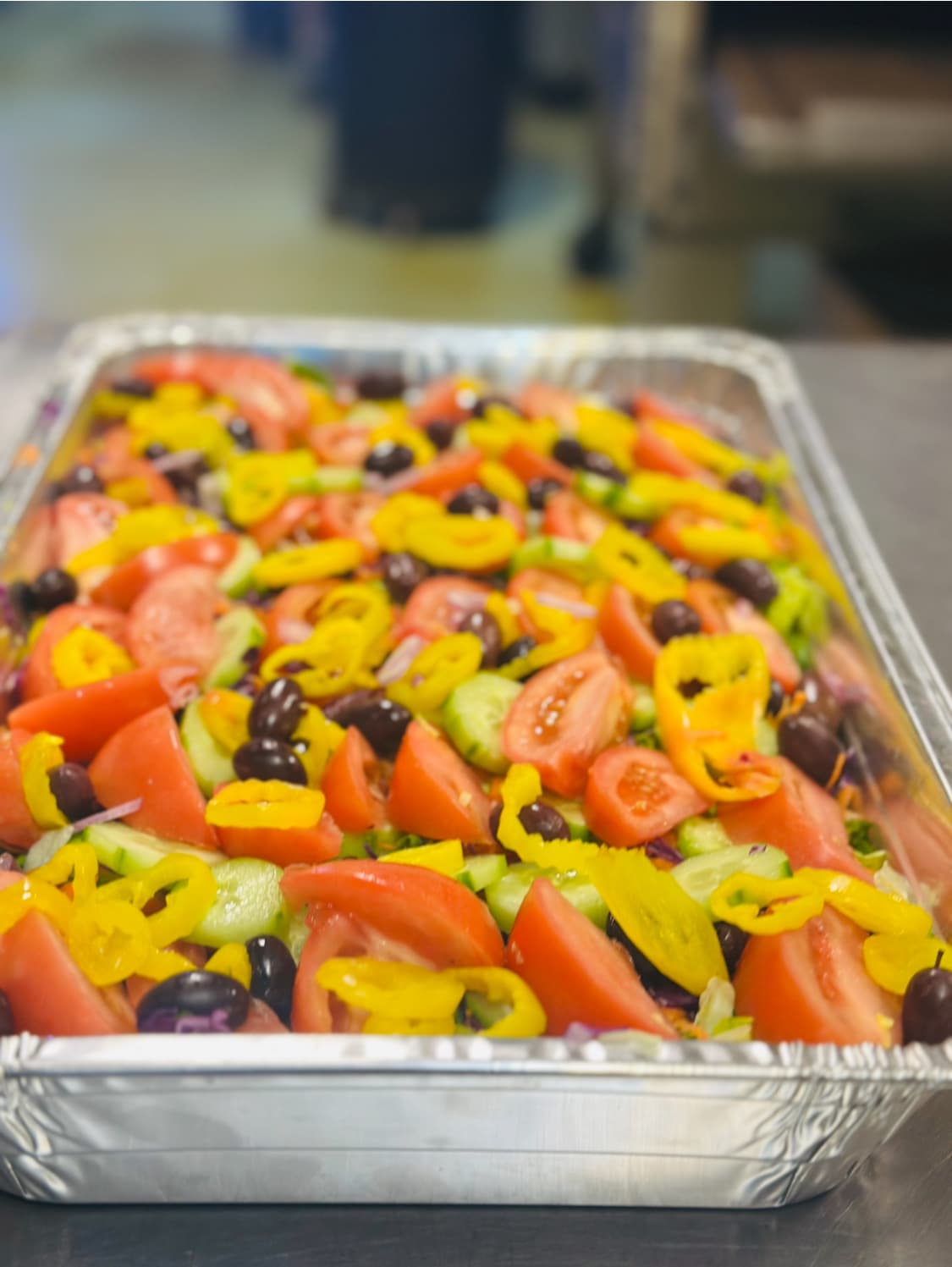 Large catering tray of Greek salad with tomatoes, cucumbers, yellow peppers, and black olives.