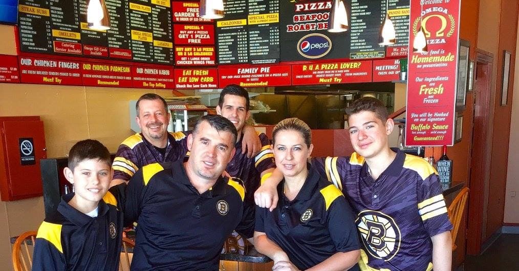 Five people in matching Boston Bruins jerseys pose in front of a pizza shop menu.