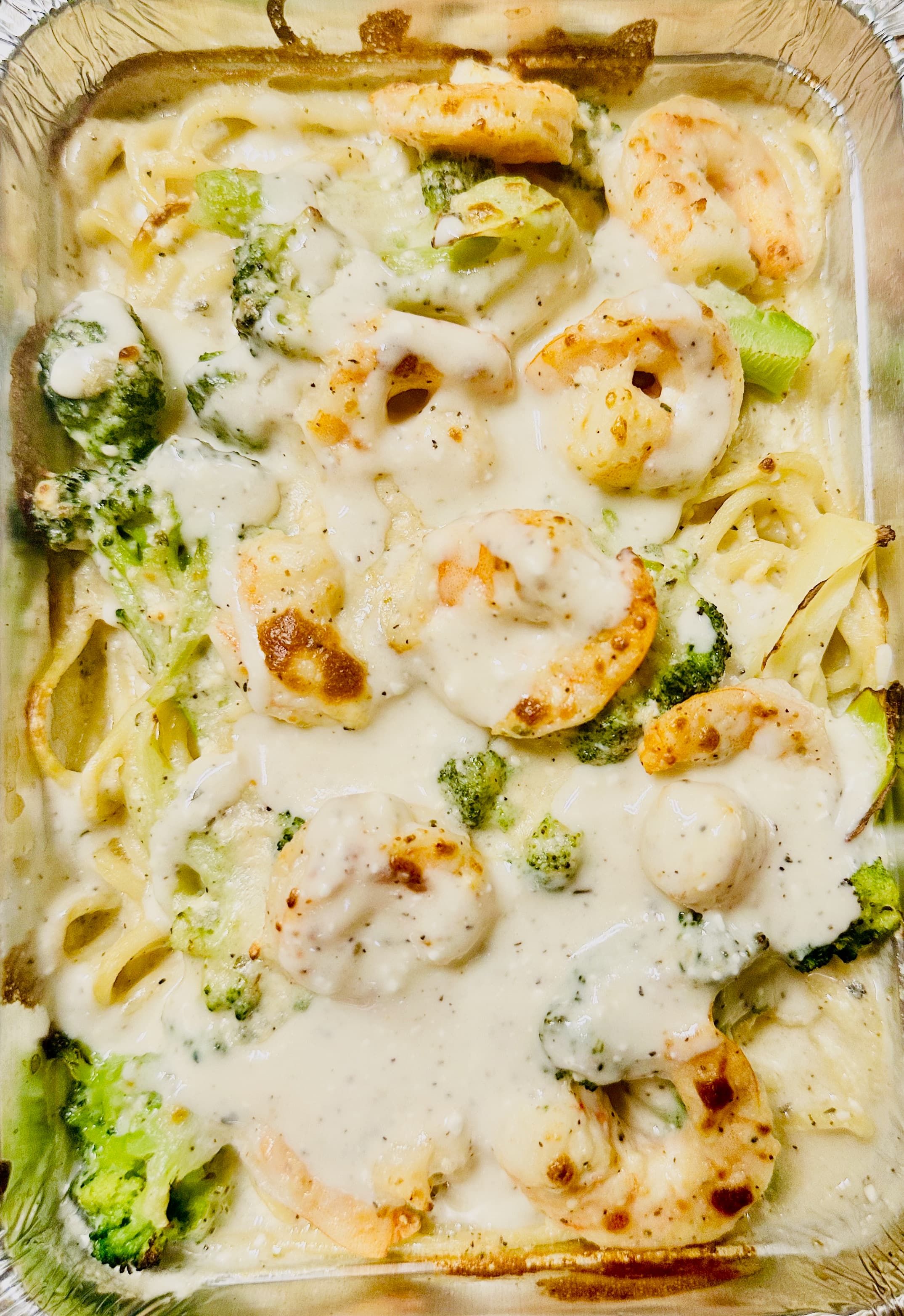 Creamy Alfredo pasta with large shrimp and broccoli florets in a disposable aluminum foil tray.